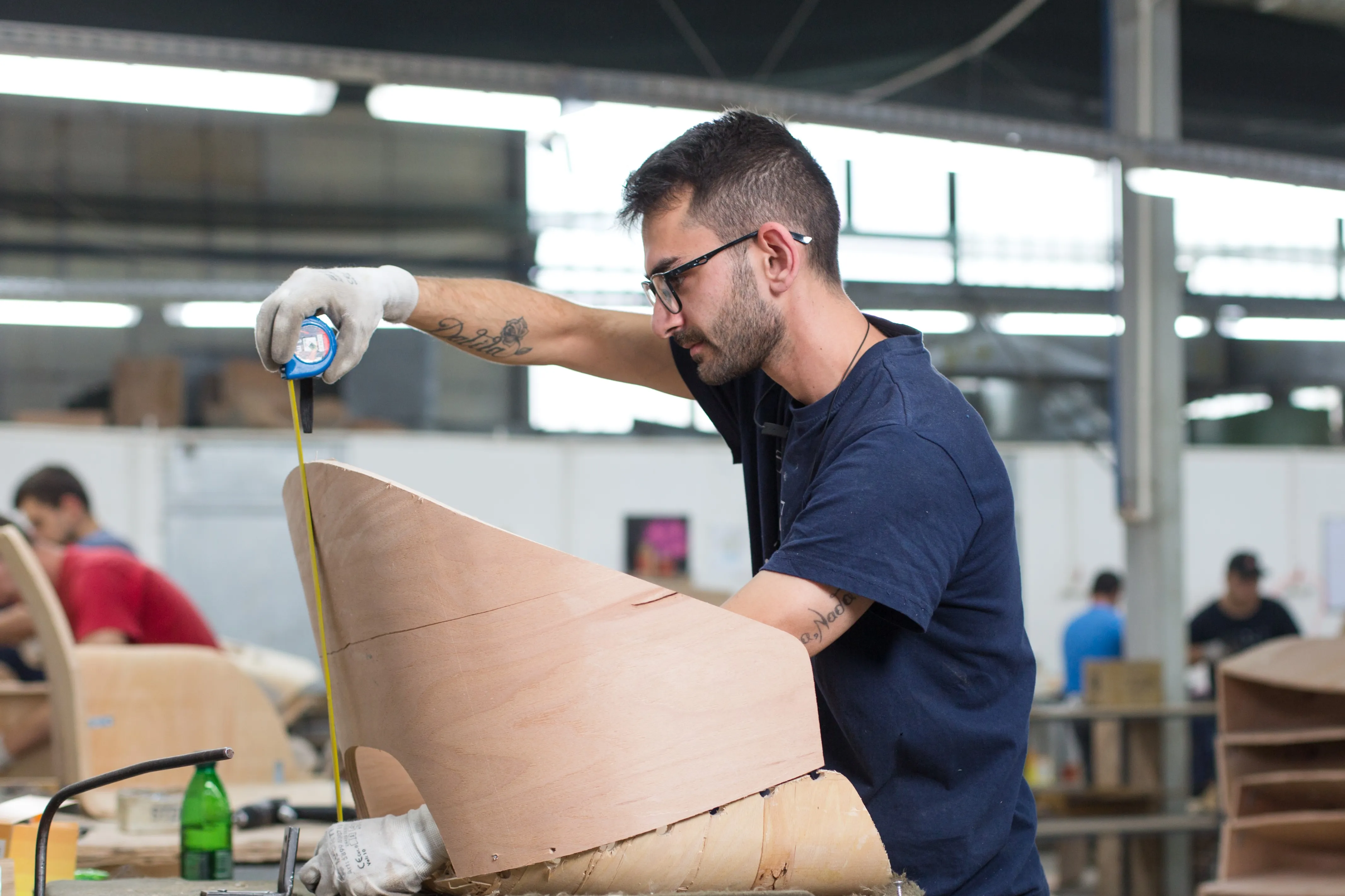 A worker checking dimensions on a curved plywood seat with a tape measure.